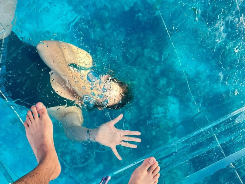 Vue spectaculaire à travers le plancher de verre montrant un baigneur nageant sous la coque du bateau transparent.