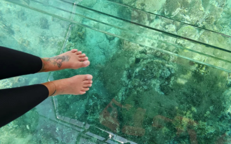 Vue en première personne des pieds sur le fond transparent du bateau, révélant les coraux et les rochers sous l'eau.