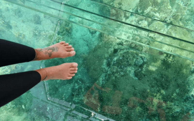 Vue en première personne des pieds sur le fond transparent du bateau, révélant les coraux et les rochers sous l'eau.